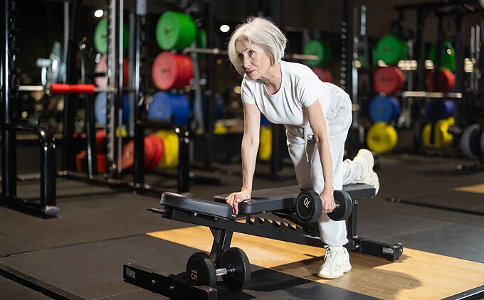 Mujer mayor realizando ejercicio de remo con mancuerna en banco plano dentro de un gimnasio