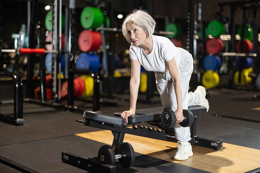Mujer mayor realizando ejercicio de remo con mancuerna en banco plano dentro de un gimnasio Mujer mayor realizando ejercicio de remo con mancuerna en banco plano dentro de un gimnasio